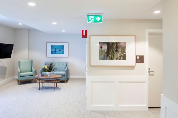 Bright, modern institutional or commercial interior reception/lounge area with pale grey walls, white panelled doors, recessed ceiling lights, and light carpet. A small seating arrangement includes a pale green armchair, blue upholstered sofa, and round timber coffee table. Framed artwork, an illuminated emergency exit sign, and a fire extinguisher sign are visible, with a partially open doorway on the right.