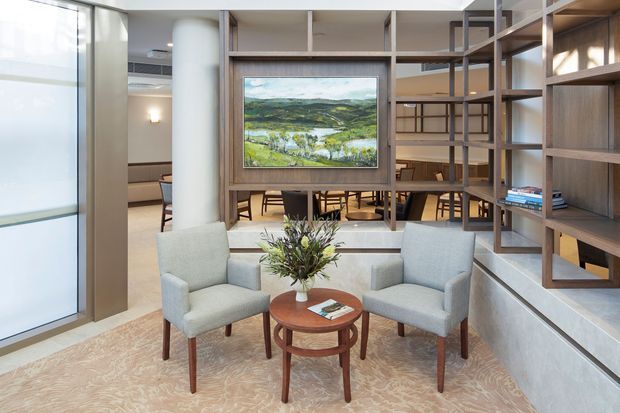 A refined communal lounge or waiting area with two upholstered armchairs arranged around a round timber coffee table, set on a patterned carpet. A large open timber shelving and room-divider structure frames a dining or meeting area behind, with a landscape artwork as a focal point. The space uses warm timber joinery, pale neutral finishes, glazing, and contemporary furnishings.