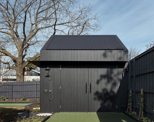 A compact dark charcoal corrugated-metal building with a steep pitched roof and vertically ribbed facade, positioned in a fenced landscaped yard. The structure features a large double-door opening or garage-style doors, exterior wall lighting, and a small artificial-turf forecourt. Mature deciduous trees and neighbouring fencing surround the building.
