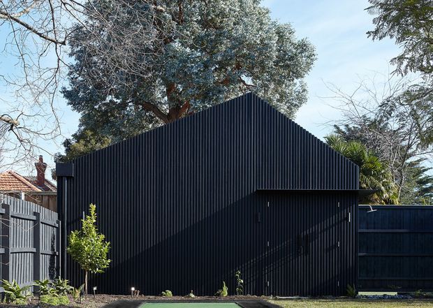 Exterior view of a dark corrugated-metal-clad structure with a simple gabled roof and large integrated garage or entry doors. The minimalist building sits behind a matching dark fence, with established trees and residential surroundings visible around it.