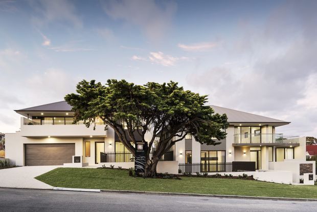 Contemporary multi-storey residential facade viewed from the street, featuring broad low-pitched roof planes, white rendered walls, extensive glazed openings, balconies, an integrated double garage, and a large sculptural evergreen tree in the front garden.