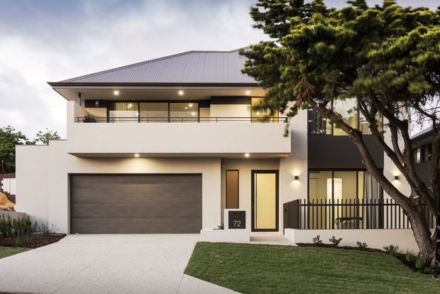A contemporary two-storey residential facade with a broad low-pitched metal roof, crisp white rendered walls, dark window and door frames, an integrated double garage, upper-level balcony, and a landscaped front lawn. Mature trees partially frame the right side of the property.