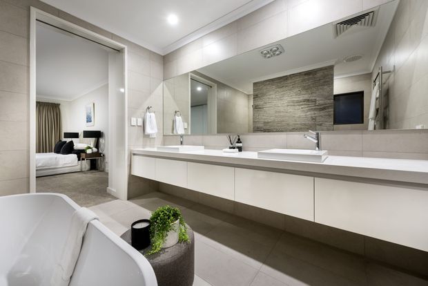 Contemporary bathroom with a long floating double vanity, two basins, extensive mirrored cabinetry, pale large-format wall and floor tiles, and a textured grey feature wall. A freestanding white bathtub occupies the foreground, while an open doorway reveals part of an adjacent bedroom.