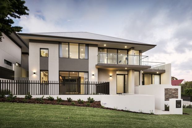 Contemporary two-storey residence with a broad low-pitched metal roof, rendered white and grey facade, extensive dark-framed glazing, glass balcony balustrade, recessed lighting, and a black vertical-bar front fence. A landscaped lawn and low white entry wall frame the street-facing exterior.