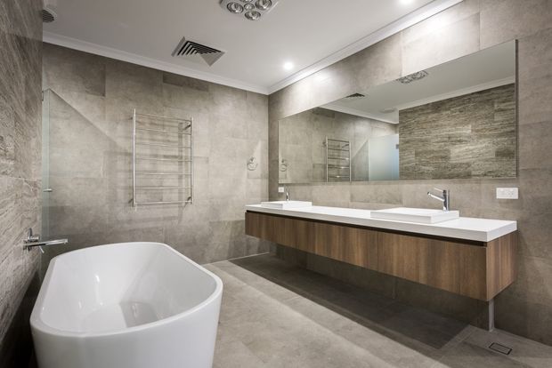 Contemporary bathroom with large-format grey stone-look wall and floor tiles, a freestanding white bathtub, floating dark timber vanity with a white benchtop and integrated basins, chrome tapware, heated towel rail, and a large wall mirror. Recessed ceiling lighting and ventilation contribute to a clean, refined fit-out.