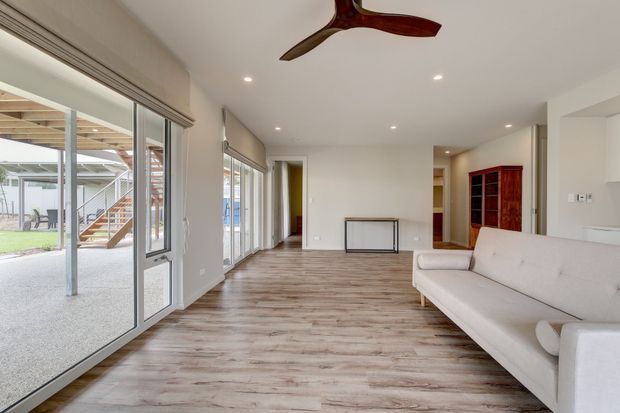 Bright, elongated open-plan living room with pale walls, recessed ceiling lights, timber-look flooring, extensive glazed doors and windows opening to a covered outdoor area, and a light upholstered sofa. A dark timber ceiling fan and built-in timber cabinetry add contrast. The room appears largely unfurnished and airy.