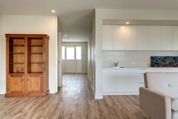 Bright open-plan interior showing a contemporary white kitchen opening to a living area and hallway. The room has pale timber-look flooring, white cabinetry, recessed ceiling lights, a tiled splashback, and a large traditional timber display cabinet along the left wall. A neutral upholstered armchair is visible in the foreground.