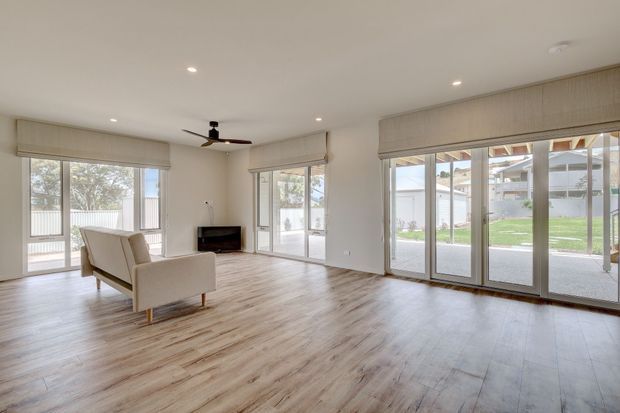 Bright, spacious open-plan living room with pale timber-look flooring, white walls, extensive floor-to-ceiling glazed doors and windows, roller blinds, recessed ceiling lights, a ceiling fan, and a simple freestanding sofa facing a dark fireplace or media unit. The room opens visually to a paved outdoor area and lawn.