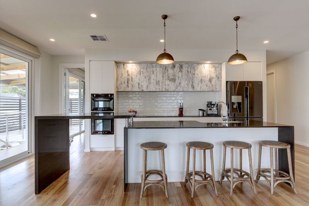 Bright contemporary kitchen with a large island and four stools, white cabinetry, a dark benchtop, textured grey-white feature cabinetry, pendant lighting, stainless-steel appliances, and timber flooring. Glazed doors at left open toward an outdoor area.
