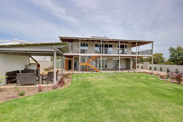 Exterior view of a raised two-storey house with broad covered balconies, horizontal metal balustrades, extensive glazing, an external stair, and a separate covered outdoor entertaining area with a built-in fireplace. The building sits within a manicured lawn and landscaped garden.