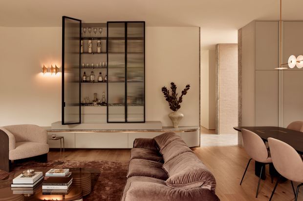 Warm, refined open-plan living and dining area with a neutral palette. The room features a large plush sectional sofa, sculptural lounge chair, dark timber coffee table, textured rug, and a built-in low console with a glass-fronted drinks display cabinet. A dining table with upholstered chairs sits to the right, while contemporary pendant and wall lighting add soft ambient illumination. The space has clean-lined cabinetry, timber flooring, and subtle decorative styling.
