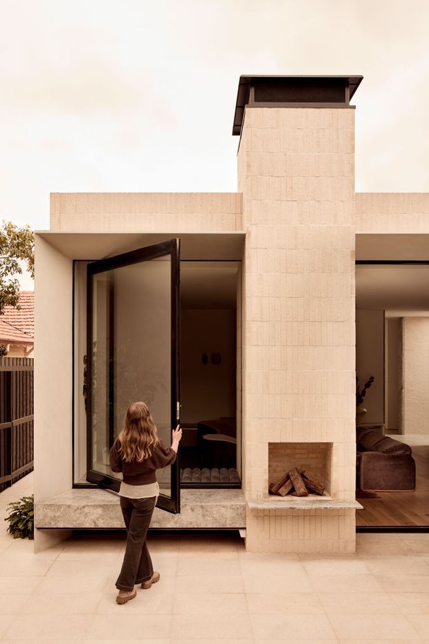 Exterior view of a contemporary residence with a pale textured masonry facade, projecting flat rooflines, a tall central chimney, and a built-in open fireplace. Large dark-framed glazed doors open onto a paved terrace, with a living room visible inside. The composition emphasizes the home's facade and indoor-outdoor connection.