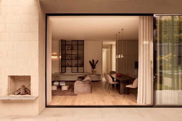 View from an outdoor terrace into a refined open-plan living and dining area through wide glazed sliding doors. The interior features pale timber flooring, warm beige walls and cabinetry, a softly upholstered sofa, dining table with upholstered chairs, a dark-framed glass display cabinet, and minimalist pendant lighting. A built-in masonry fireplace is visible on the exterior wall at left, with sheer curtains and garden views at right.