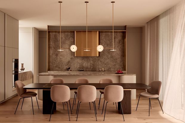 A refined open-plan dining and kitchen space in warm neutral tones. A long dark timber dining table with upholstered dining chairs occupies the foreground, while the rear kitchen features full-height cabinetry, a central island, a dramatic stone-look feature wall, timber cabinetry, and pendant lighting. Sheer floor-to-ceiling curtains soften the right side, and timber flooring runs throughout.