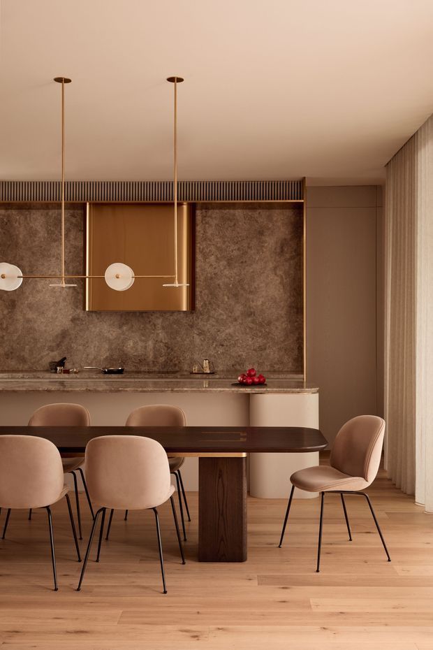 Warm, refined open-plan dining and kitchen interior with a long dark timber dining table, upholstered dining chairs, a compact kitchen island, and a richly textured brown feature wall. The space features light timber flooring, bronze-toned fixed cabinetry or wall paneling, slim sculptural pendant lights, vertical timber screening, and full-height sheer curtains.