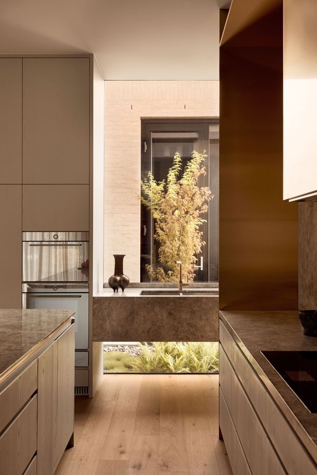 A warm, contemporary galley-style kitchen with full-height taupe cabinetry, integrated stainless-steel appliances, stone benchtops, and timber flooring. A central island occupies the foreground, while a dark stone or textured splashback zone and sink sit beneath a tall dark-framed window. A low glazed opening at floor level provides a view to planted greenery, adding natural light and an indoor-outdoor connection. The palette is earthy and refined, with timber, bronze-brown cabinetry accents, and soft neutral walls.