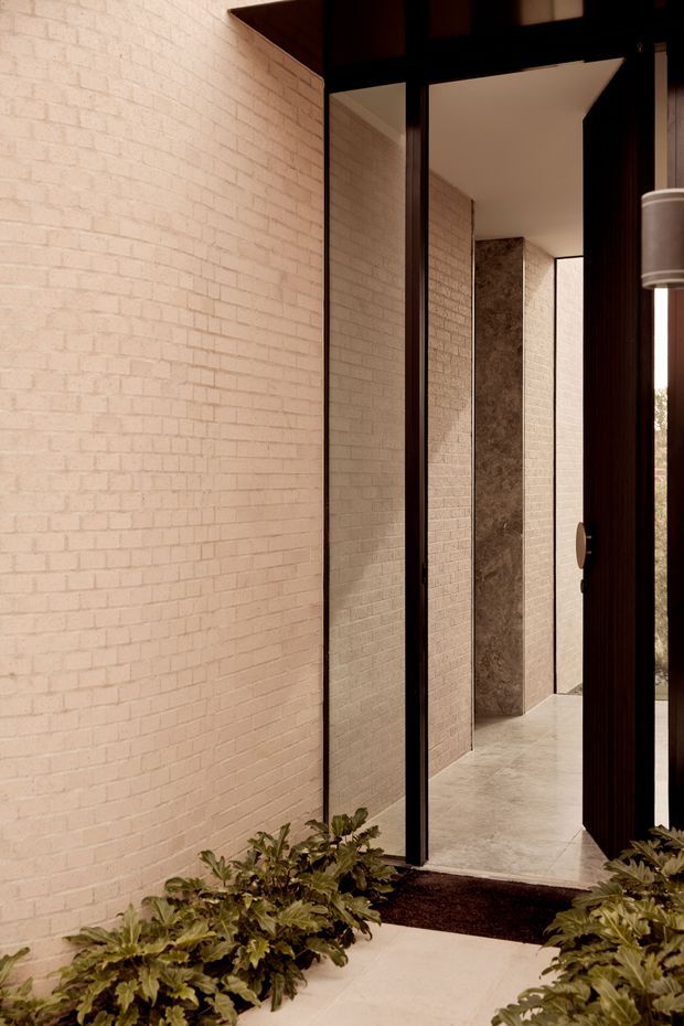 A refined residential entryway viewed from outside, with a tall dark timber-framed pivot door and narrow glazed side panels opening into a minimalist corridor. Pale textured brick walls continue through the entry, while large stone-clad vertical panels and light stone paving create a warm, tonal material palette. Low leafy planting borders the path, and a dark entry mat sits at the threshold. The composition emphasises strong vertical lines, deep reveals, and framed views into the home.