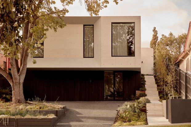 Front exterior view of a contemporary two-storey residence with a minimalist rectilinear form, pale rendered or masonry upper facade, dark timber-clad ground level, tall black-framed windows, recessed entry, and an integrated dark garage opening. A landscaped front garden, driveway, and steps lead to the entry, with neighbouring homes visible at the sides.
