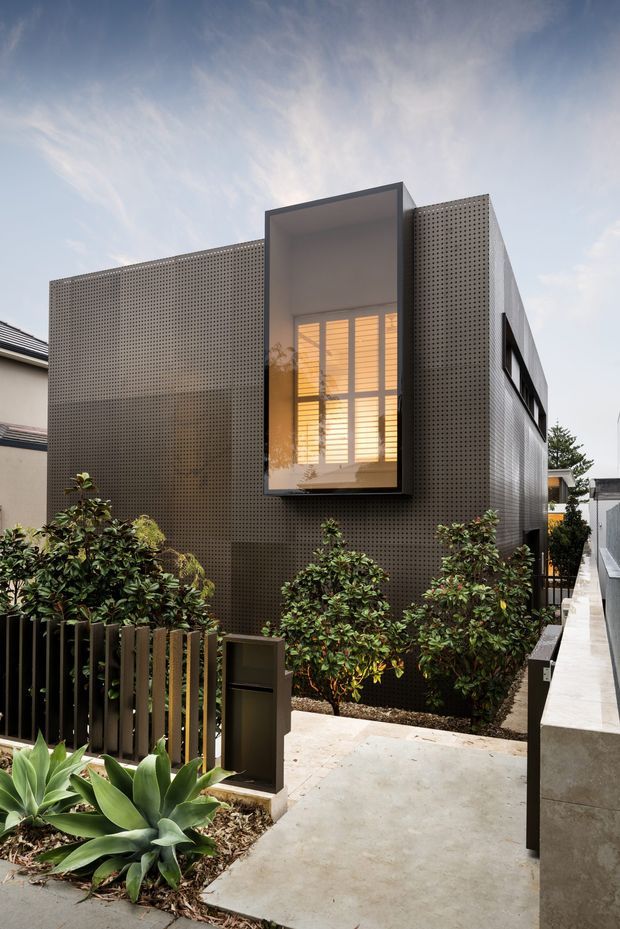 Exterior view of a compact contemporary multi-level residence, featuring dark perforated facade panels, a projecting recessed window bay with warm interior lighting, and a narrow landscaped entry path with dense shrubs and agave planting.