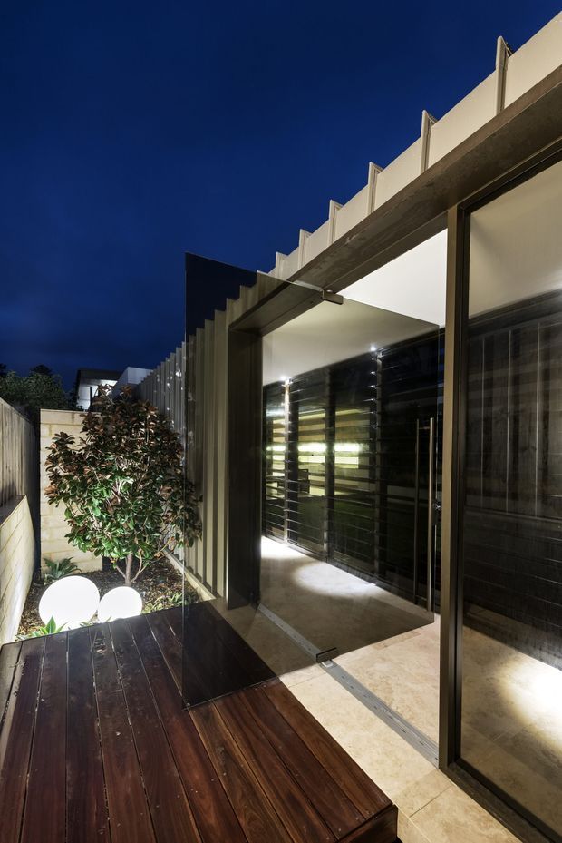 Night-time view of a contemporary residential exterior and entry terrace. A dark timber deck leads alongside a landscaped garden and illuminated spherical lights, toward a covered passage with large glazed sliding doors, dark metal framing, and a projecting concrete canopy with vertical fins. The scene is strongly lit against a deep blue sky.