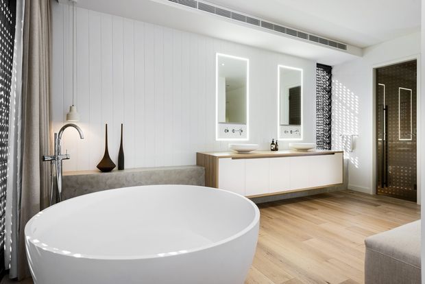 Bright contemporary bathroom featuring a freestanding white oval bathtub, double vanity with illuminated mirrors, timber-look flooring, vertical white wall panelling, and a glass-enclosed shower with dark patterned tile. Natural light filters through patterned privacy screens, creating a clean, refined atmosphere.