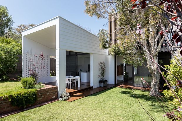 A bright contemporary garden-facing residential extension with white horizontal cladding, a deep open-sided covered terrace, large glazed openings, and dark timber decking. The extension connects to a taller darker timber-clad building behind it, opening onto a manicured lawn with brick-edged garden beds and mature planting.