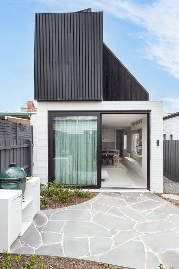 Rear exterior view of a compact contemporary home extension, featuring a white-painted brick lower facade, dark vertical timber cladding and a steep gabled upper form. Large black-framed glazed doors open onto an irregular natural-stone paved courtyard. A built-in outdoor kitchen or barbecue area is visible at left, with the interior kitchen and living area seen through the glazing.