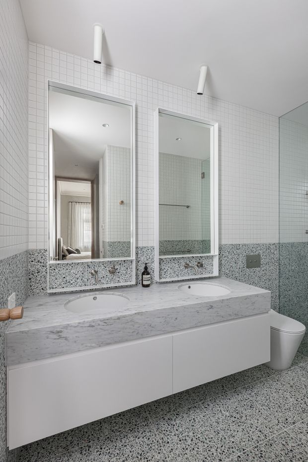A bright contemporary bathroom featuring a double floating vanity with integrated basins, a pale marble-look benchtop, white cabinetry, twin rectangular mirrors, and wall-mounted cylindrical lights. Small white square wall tiles cover the upper walls, while grey terrazzo-style tiles create a contrasting lower wall band and floor. A toilet and large wall mirror are visible at the right.