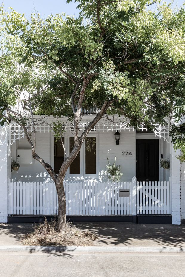 Street-facing exterior of a compact white-painted brick residence with a black entry door, white picket fence and gate, decorative white metal verandah trim, front windows, mailbox, and mature street trees. The facade has a restrained heritage-inspired character with strong black-and-white contrast.