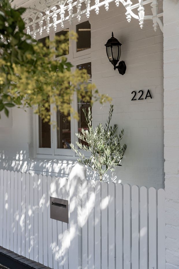 Close-up of a refined white-painted residential entry facade. The brick wall features a black traditional lantern, black house number 22A, glazed entry doors, ornate decorative white fretwork under the porch roof, and a white picket fence with a metal letterbox. An olive tree and leafy planting soften the entrance.