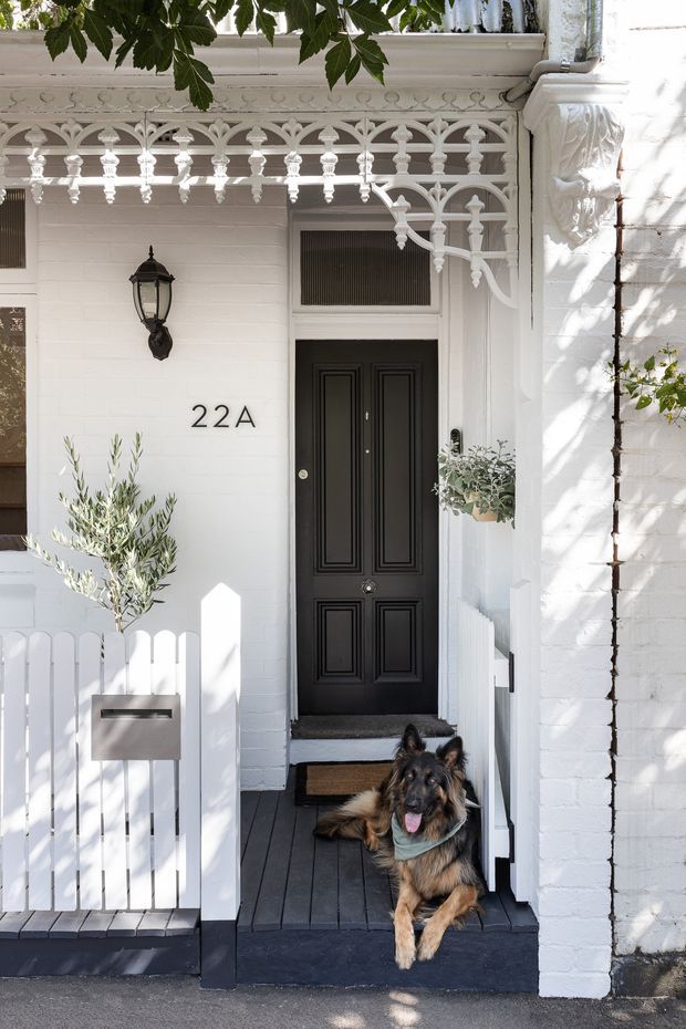 Bright exterior view of a white-painted heritage townhouse entry. The facade features ornate decorative fretwork, a black panelled front door, traditional wall lantern, white picket gate and fence, house number 22A, dark timber decking and steps, and a small hanging planter. A dog sits on the front steps. The image highlights the decorative entry facade and period detailing.
