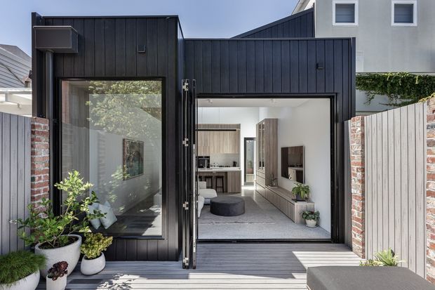 Exterior view of a contemporary narrow home extension or entry facade, clad in dark vertically profiled panels with a large glazed opening and open pivot door. A timber deck and potted plants lead into a bright living area, where built-in timber cabinetry, a television wall and kitchen joinery are visible. Brick boundary walls and a pale vertical screen fence frame the entry.