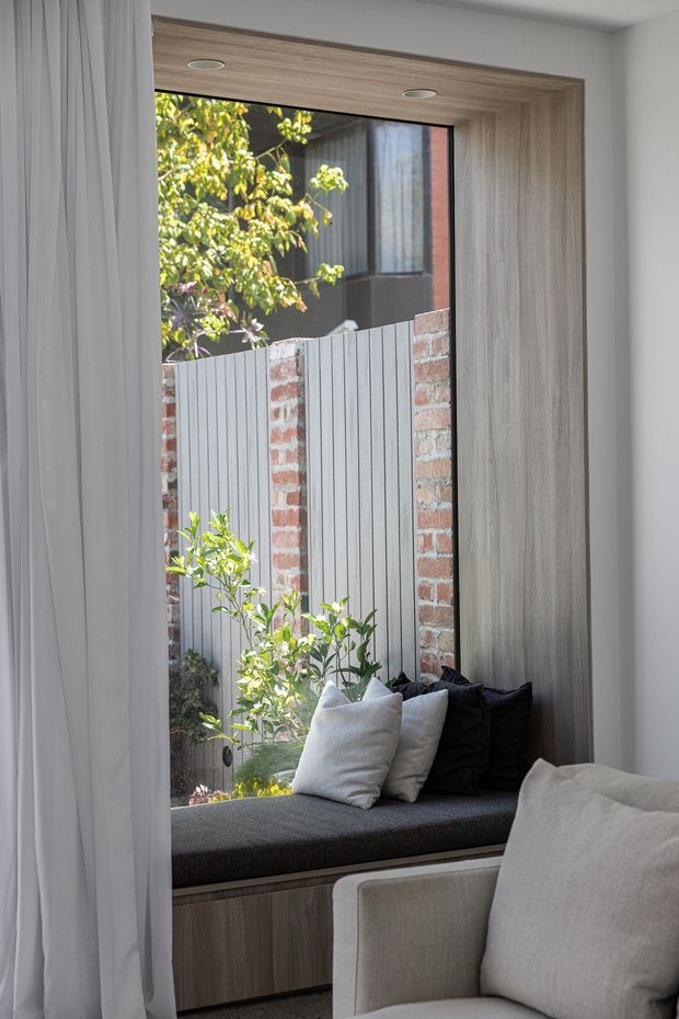 A refined interior corner featuring a built-in window seat beneath a large floor-to-ceiling window. The seat has dark charcoal upholstery, light and black cushions, and timber-grain cabinetry below. Full-height white curtains frame the window, while a pale upholstered armchair occupies the foreground. Outside, a brick-and-grey-panel boundary fence and leafy planting are visible.