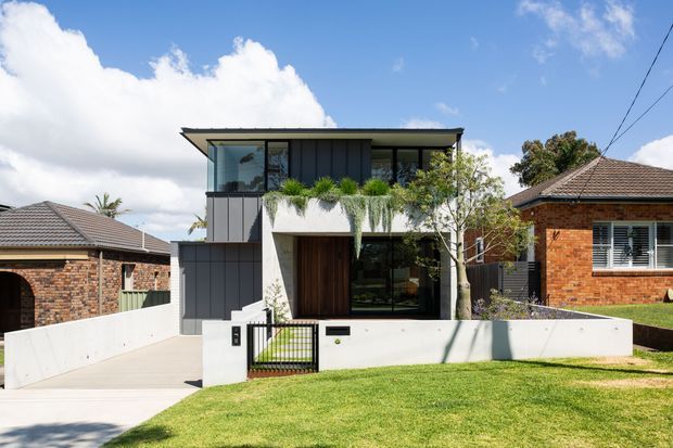 Street-facing view of a contemporary two-storey residence with a low-pitched roof, dark vertical cladding, extensive glazing, a recessed timber entry door, and a white rendered balcony planter. The home is set behind a low white boundary wall and landscaped lawn, with neighbouring brick houses visible on either side.