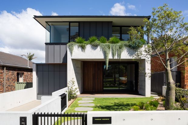 Front exterior view of a contemporary two-storey residence with a broad low-pitched roof, dark vertical cladding, concrete entry portal, extensive balcony planting, timber front door, and a small landscaped lawn behind a low boundary wall and gate.