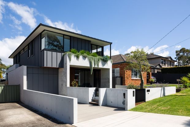 Street-facing view of a contemporary two-storey residence with dark vertical cladding, extensive upper-level glazing, a projecting flat roof, and exposed concrete entry walls and boundary walls. The home sits in a suburban streetscape beside established brick and rendered houses, with a driveway, front lawn, and cascading greenery over the balcony edge.