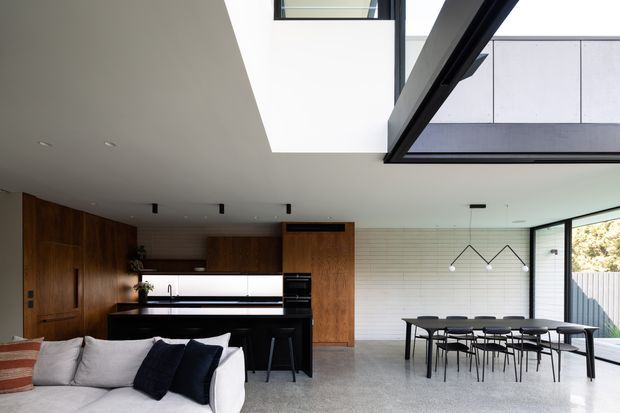 Open-plan contemporary kitchen, dining, and living area with a dramatic double-height void and clerestory glazing. The kitchen features dark timber wall cabinetry, a white island with dark base cabinetry, and integrated appliances. A dining table with black chairs sits beside a white horizontally tiled feature wall, while full-height glazing opens toward a fenced outdoor area. The space has polished concrete flooring, restrained black lighting, and a neutral monochrome palette with warm timber accents.