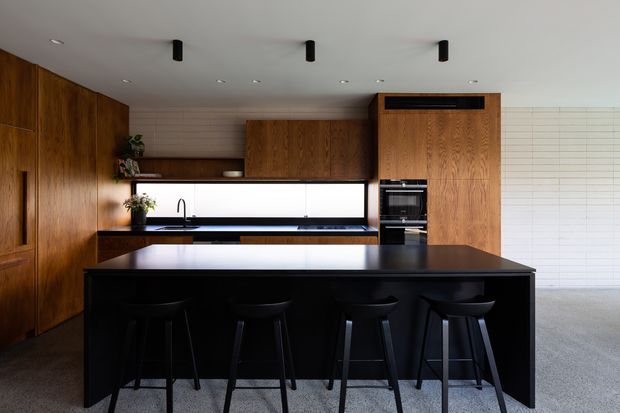 Modern open-plan kitchen dominated by a large black island with four bar stools. Warm timber cabinetry runs along the left and rear walls, with integrated appliances, a black benchtop, a dark sink and tap, and a long horizontal window splashback. White rectangular wall tiling and a pale carpeted adjacent area create contrast, while minimalist black ceiling spotlights complete the restrained design.