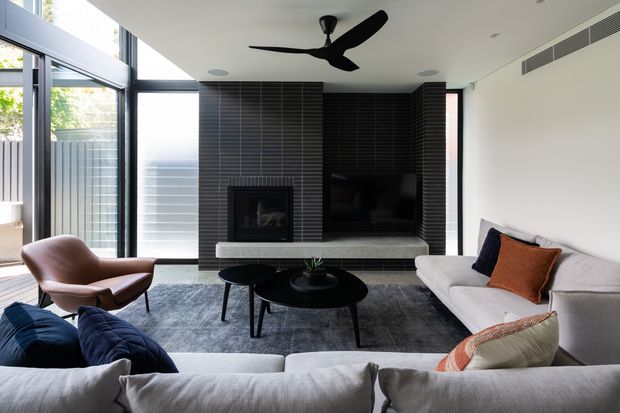 Contemporary living room with a large pale sectional sofa, sculptural brown leather lounge chair, layered round black coffee tables, and a dark grey rug. A black tiled feature wall incorporates a fireplace and television, while extensive dark-framed glazing brings in daylight. A black ceiling fan and recessed linear air-conditioning add to the clean modern fit-out.