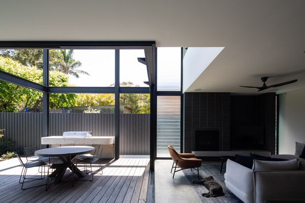A contemporary open-plan living room opening onto a timber deck and outdoor dining area through extensive black-framed glazing. The interior features a dark tiled fireplace wall with a large television, a ceiling fan, a neutral sectional sofa, and a leather lounge chair. Bright natural light and garden greenery are visible outside.
