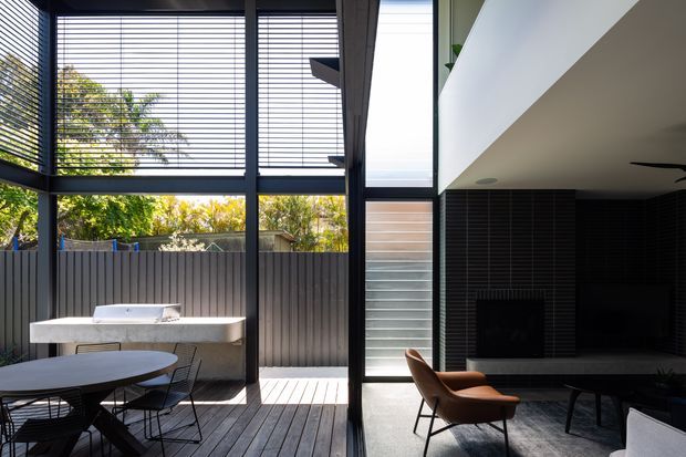 A contemporary indoor-outdoor living and dining area with floor-to-ceiling black-framed glazing and horizontal blinds. A timber deck with an outdoor dining table, chairs, and built-in barbecue or outdoor kitchen occupies the left side, while the interior on the right features a dark tiled fireplace wall, lounge seating, and a low coffee table beneath a white ceiling overhang.