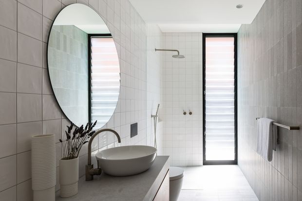 Bright contemporary bathroom with large-format grey tiled walls and floor, a floating vanity with a round vessel basin and brushed bronze tapware, an oval wall mirror, wall-mounted towel rail, toilet, and a frameless glass shower area. Narrow vertical windows with translucent blinds provide filtered natural light.