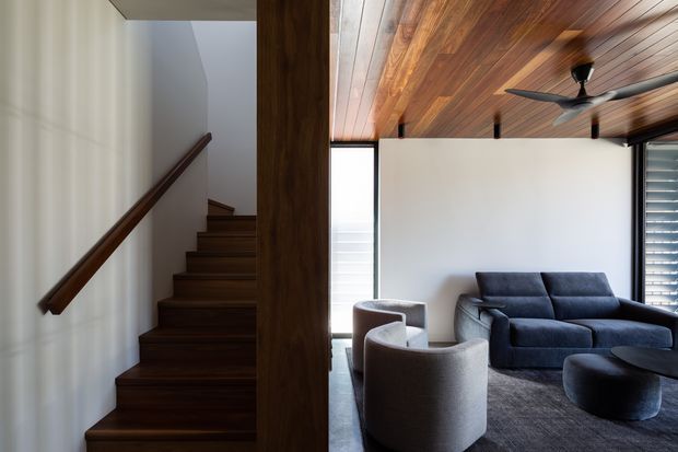 Interior view combining a dark timber stairway and a compact living room. The living area features a dark upholstered sofa, rounded armchairs, a low coffee table, large windows with louvred shading, and a striking timber-lined ceiling with a ceiling fan. White walls and dark timber finishes create a refined, warm contemporary atmosphere.