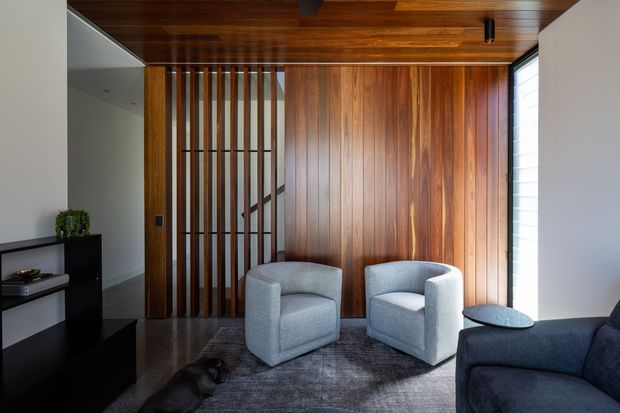 Contemporary interior living area with two light-grey upholstered lounge chairs facing a dark sofa, set on a grey rug. A rich timber-lined ceiling and full-height timber feature wall dominate the room, with a vertical timber slat screen revealing a staircase behind. Dark built-in cabinetry, polished dark flooring, and a narrow window provide contrast. The space is refined and warm with strong natural timber detailing.