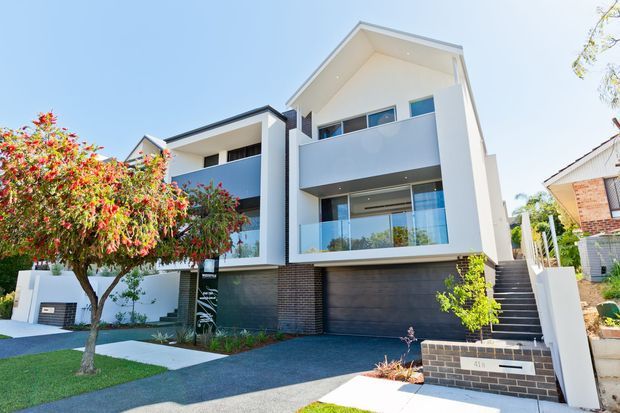 Bright exterior view of a contemporary multi-storey residence with white rendered walls, dark brick detailing, broad balconies with glass balustrades, large windows, and an integrated double garage. The home sits on a landscaped suburban site with a driveway, lawn, and mature flowering tree.