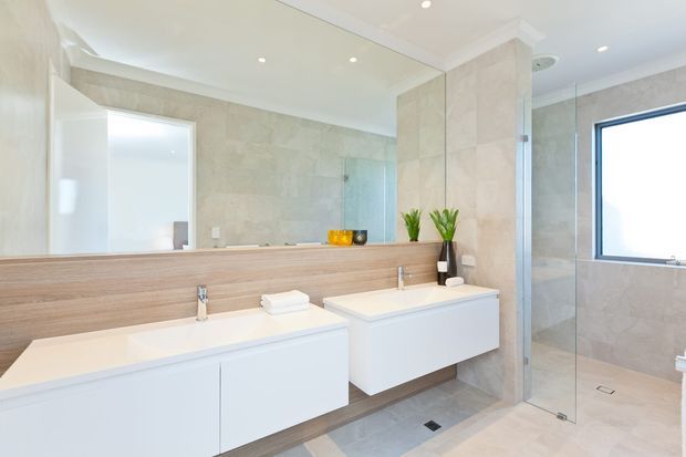 Bright contemporary bathroom with twin floating white vanities, pale timber-look backsplash, large wall mirror, beige stone-look wall and floor finishes, and a frameless glass shower enclosure. Recessed ceiling lights and minimal fittings create a clean, spacious appearance.