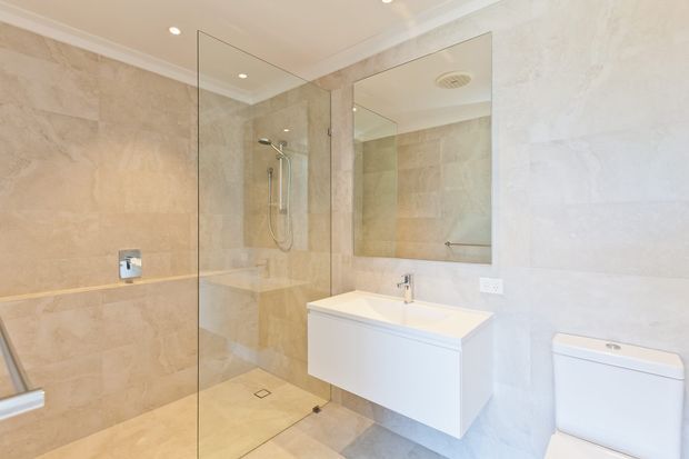 Bright contemporary bathroom featuring beige stone-look wall and floor tiles, a frameless glass walk-in shower, wall-mounted white vanity with integrated basin, large mirror, chrome fixtures, and a white toilet. Recessed ceiling lights and a clean, minimal fit-out create an airy, refined atmosphere.