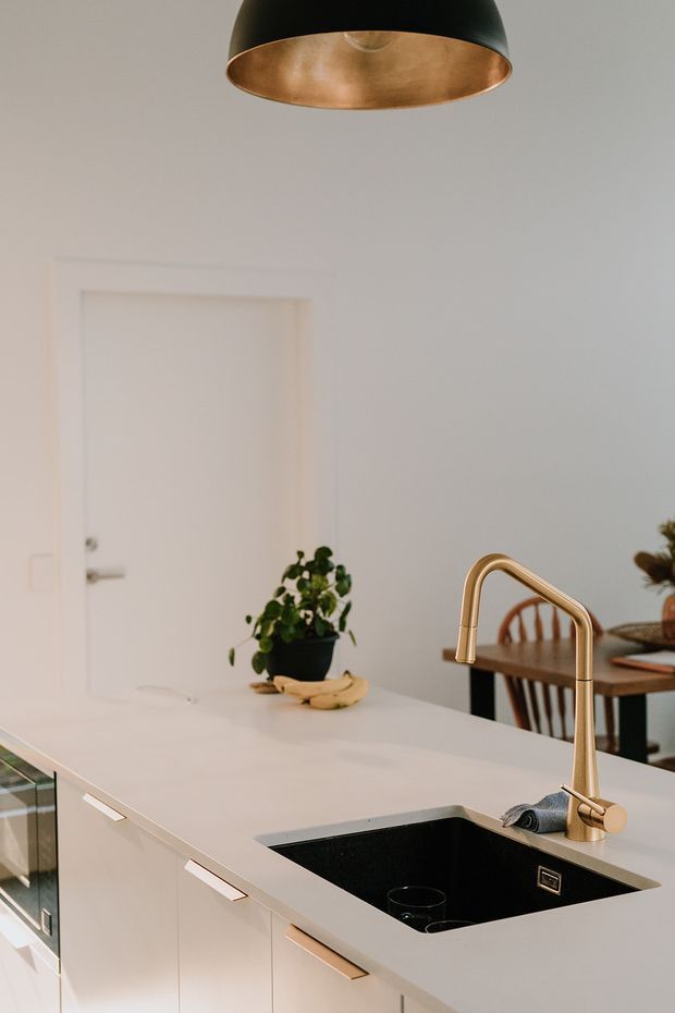 A bright contemporary kitchen viewed across a white island benchtop, featuring a deep black undermount sink, a tall brushed brass/gold gooseneck tap, white flat-panel cabinetry with slim metal handles, and a black-and-brass pendant light. A dining area and potted plant are visible in the background.