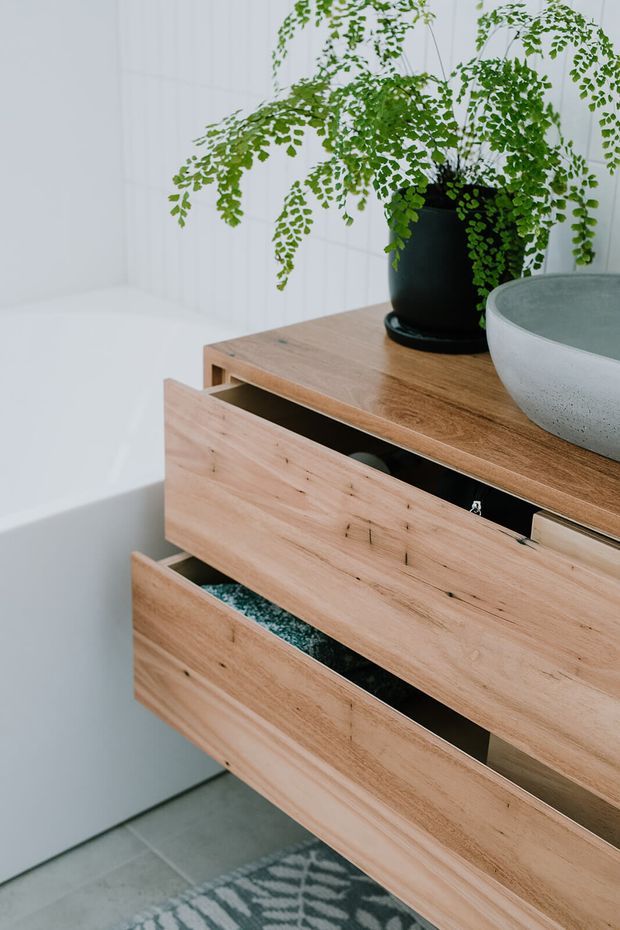 Close-up interior detail of a light timber floating vanity or storage unit with multiple open drawers, paired with a dark planter and green fern and a round grey vessel on top. The unit is set against white vertical wall panelling and grey tiled flooring, with a patterned rug partially visible below. The image has a clean, contemporary and natural feel.