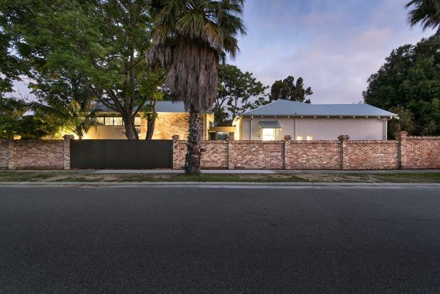 Street-level exterior view of a single-storey residential property behind a low red-brown brick boundary wall and dark metal gates. The home has a simple low-pitched hipped metal roof, light rendered walls, warm exterior lighting, mature trees, and prominent palm trees along the frontage.